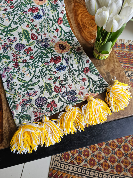 Floral-patterned table runner with yellow tassels on a wooden table.