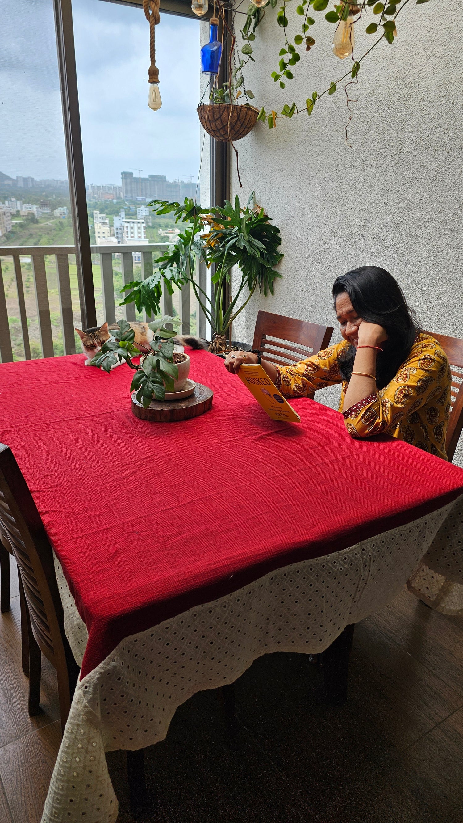 Woman sitting at a table with a red tablecloth, surrounded by plants and a window view.