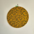 A round embroidery hoop with mustard yellow and dark green leaf motifs, framed in a wooden ring, hung on a white wall.