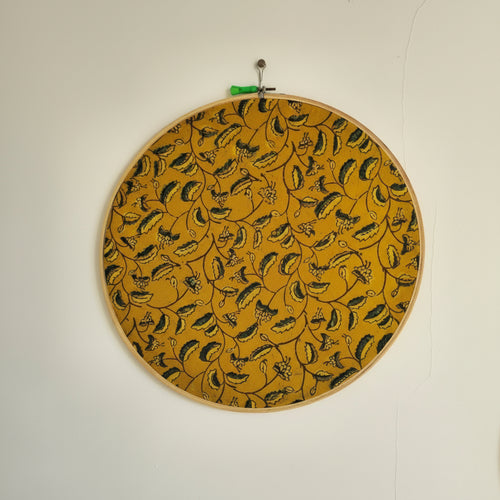 A round embroidery hoop with mustard yellow and dark green leaf motifs, framed in a wooden ring, hung on a white wall.
