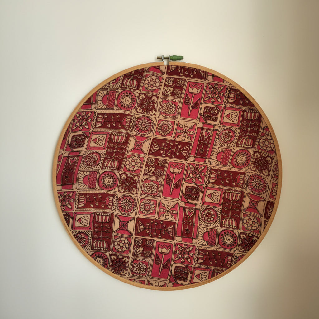 A round embroidery hoop with a geometric print Ajrakh design in abstract shades of red, mounted on a wooden frame with a visible ring at the top.