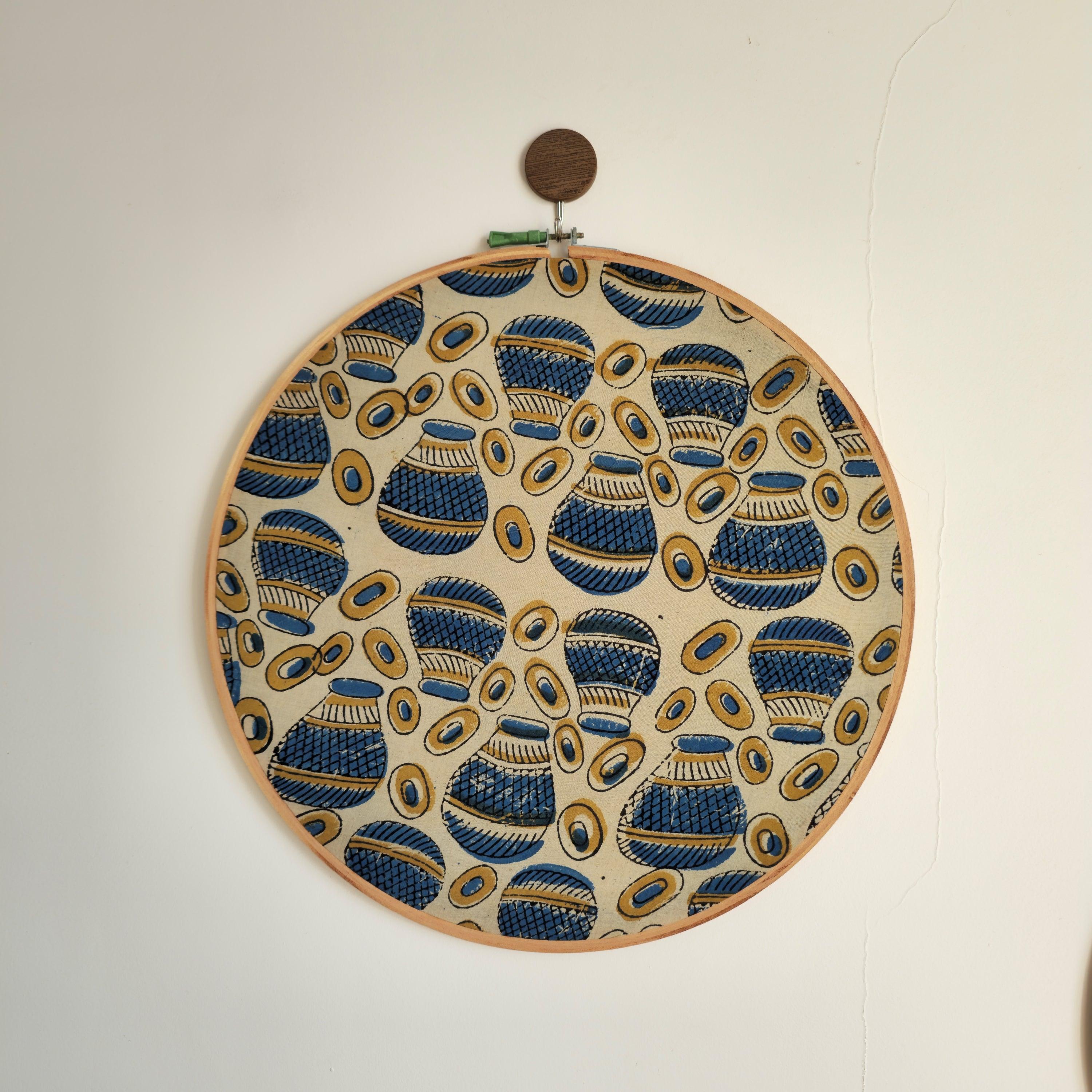A beige, indigo, and mud yellow Ajrakh block printed embroidery hoop with a quirky Matka design, framed in a wooden ring, displayed on a white wall.