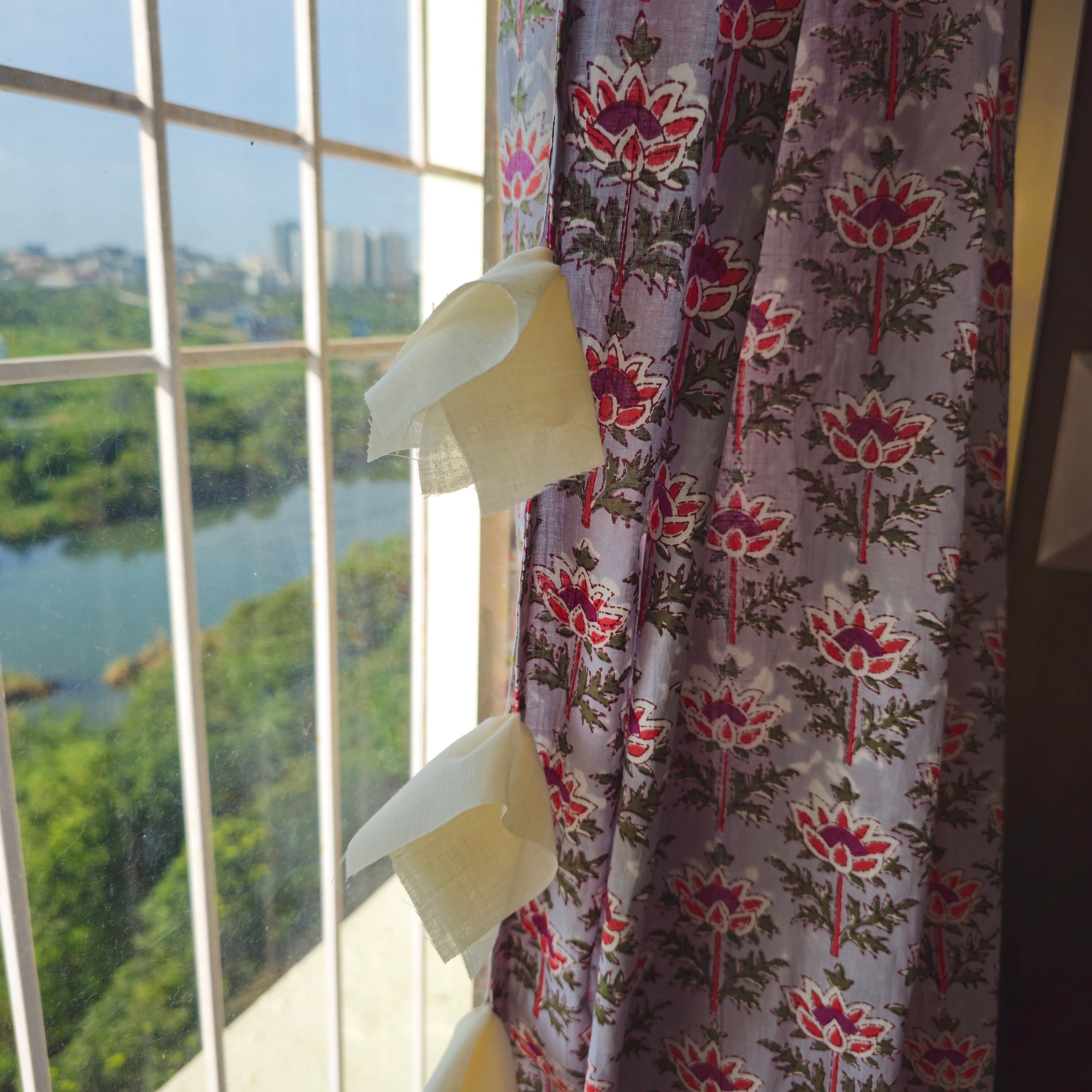 A section of a grey curtain with purple and red lotus motifs, featuring white fabric tassels, hanging in front of a window with a view of a cityscape and a river.