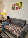 A woman sits on a gray sofa in a living room, reading a book. The sofa has a patterned throw blanket draped over the seat.
