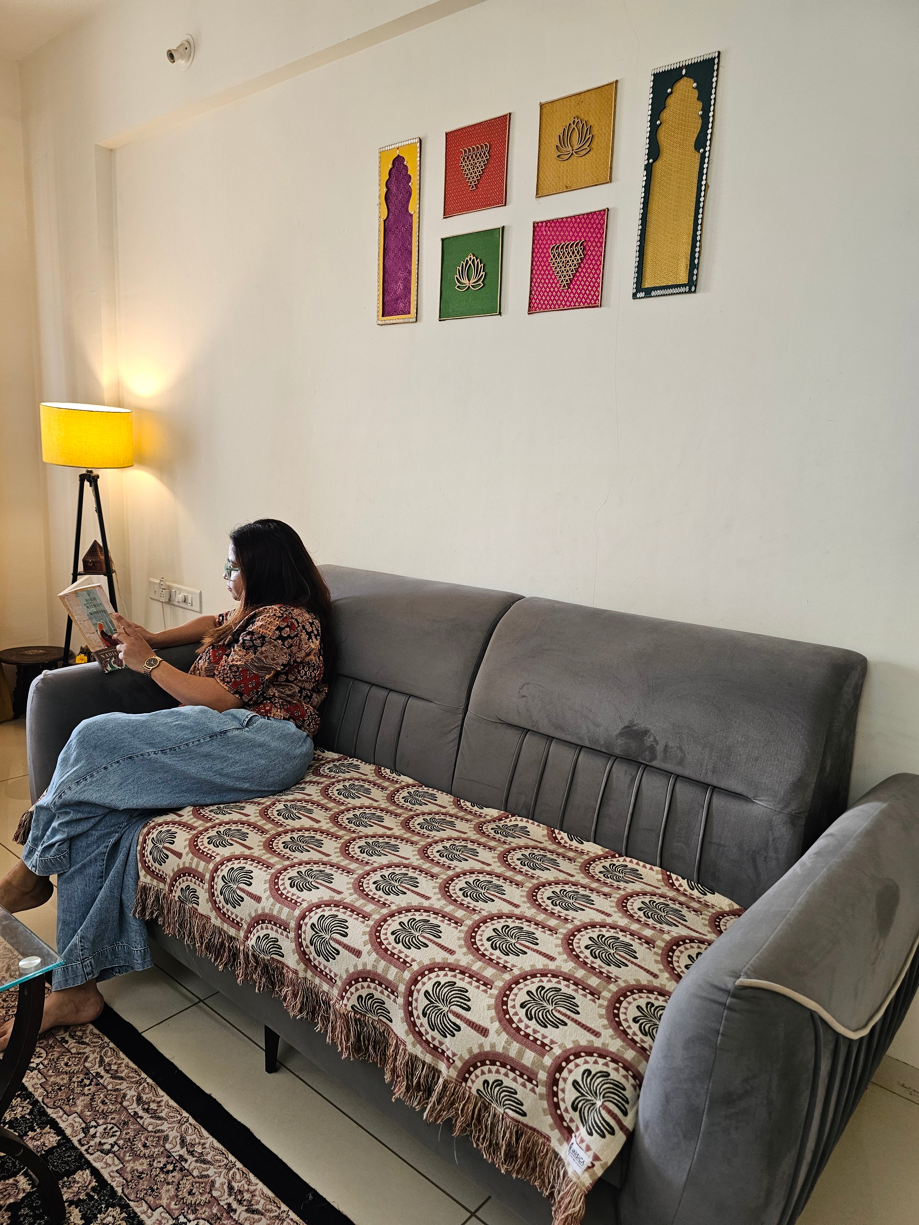 A woman sits on a gray sofa in a living room, reading a book. The sofa has a patterned throw blanket draped over the seat.