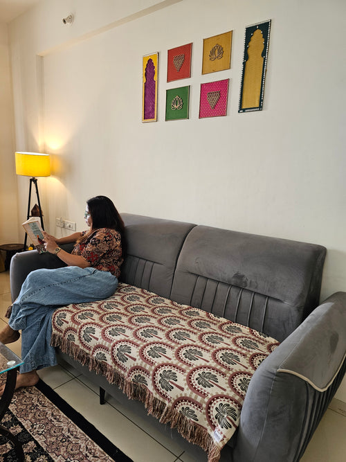 A woman sits on a gray sofa in a living room, reading a book. The sofa has a patterned throw blanket draped over the seat.