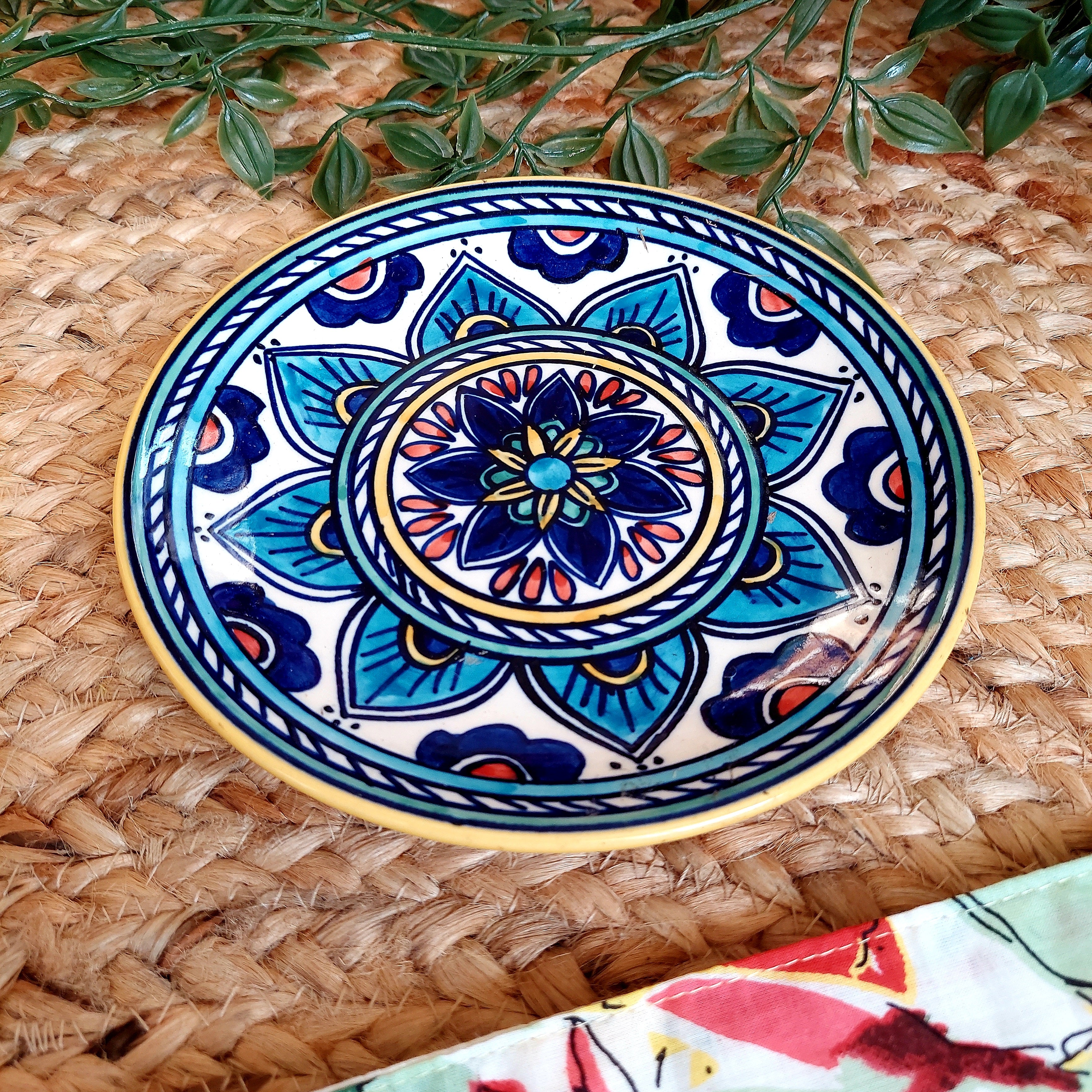A handpainted terracotta decorative wall plate with a floral and geometric pattern in blue, white, and multicolored hues, displayed on a woven basket.