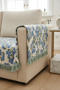 A close-up of a woven textile with a blue and green floral pattern on a white background, draped over a black chair with fringe along the bottom edge.