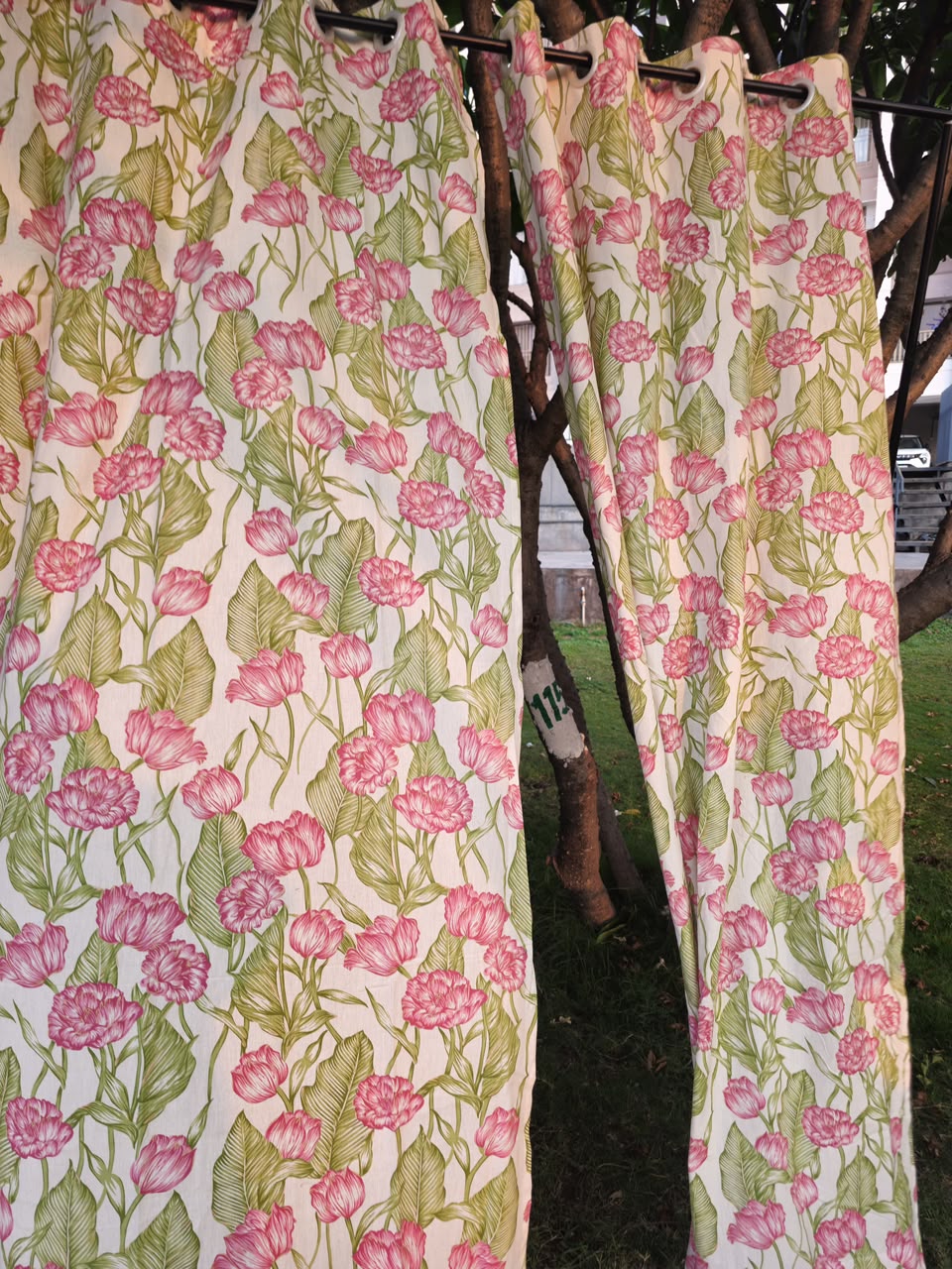 A pair of floral curtain panels hanging on a clothesline outdoors, featuring a pink and green rose pattern on a cream background.