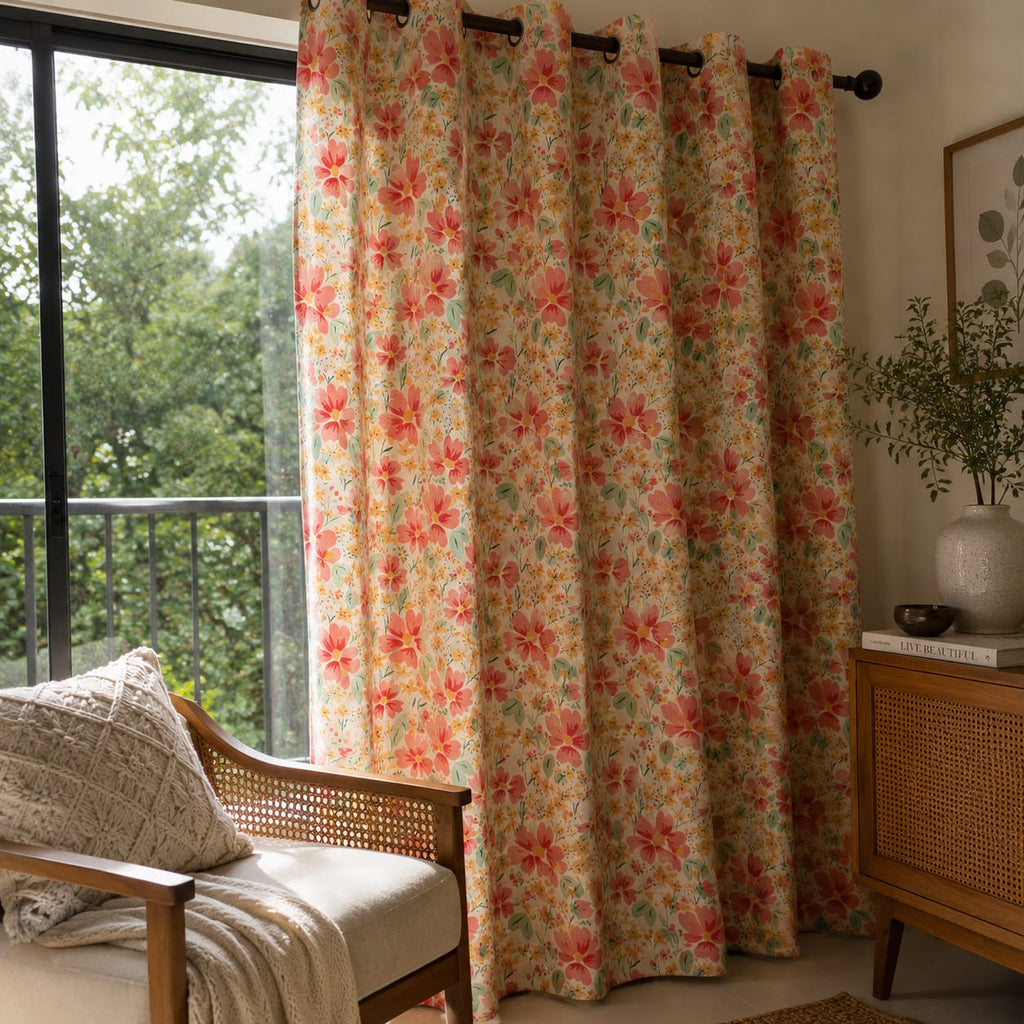 A pair of grommet-top curtains with a retro floral pattern in coral pink, orange, and green on a warm beige background, hanging in a sunlit room next to a window with a wooden chair and side table.