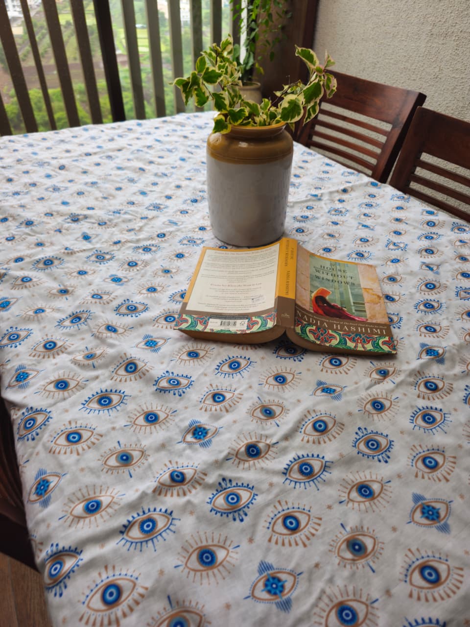 A white tablecloth with a repeating blue and beige evil eye pattern, spread across a dining table. A gray ceramic vase with a green plant and an open book are placed on the table.
