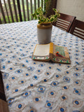 A white tablecloth with a repeating blue and beige evil eye pattern, spread across a dining table. A gray ceramic vase with a green plant and an open book are placed on the table.