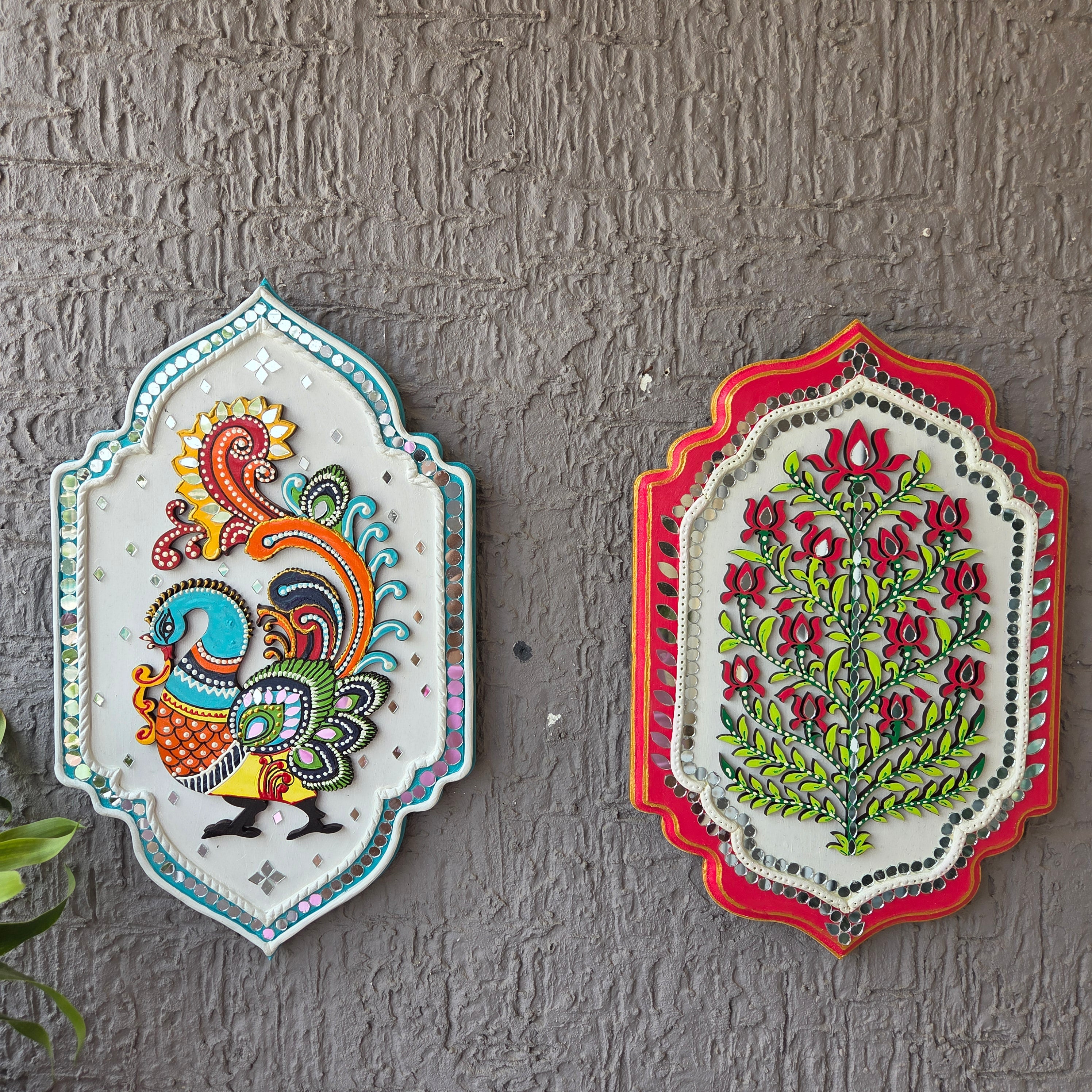 Two decorative wall art pieces with floral and peacock designs on a textured gray wall.