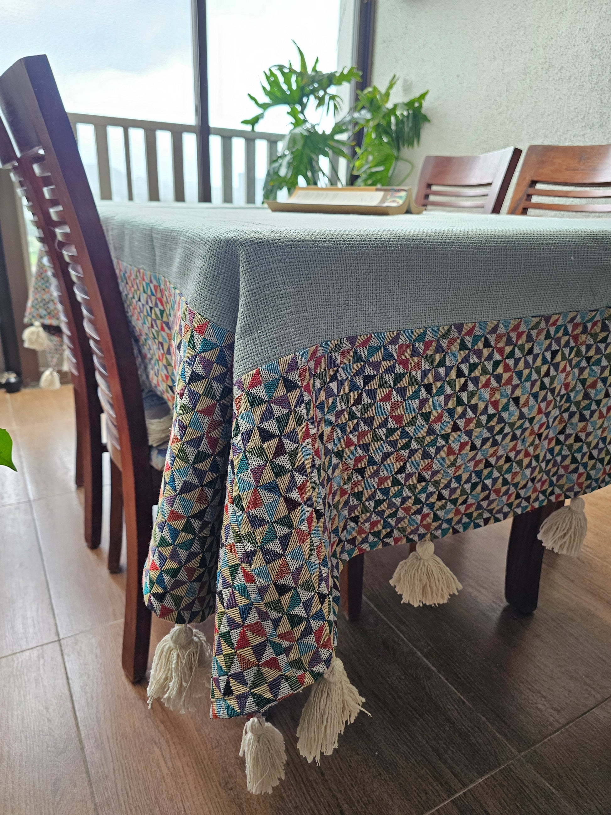 Patterned chair cover draped over a wooden chair with tassels, in a room with a window and plant.