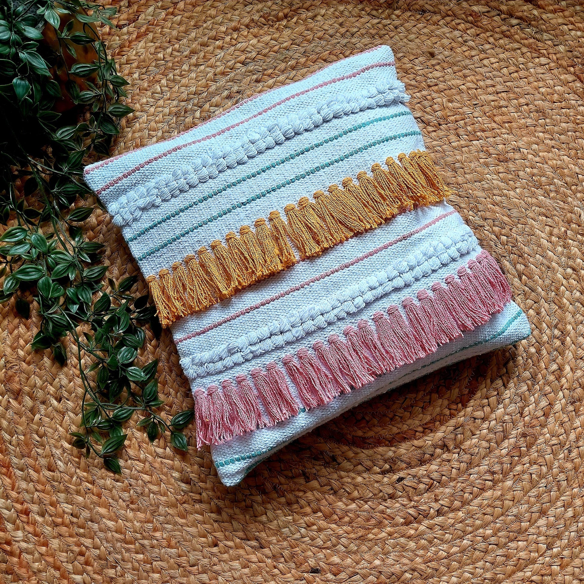Handwoven white cushion cover with pink and mustard yellow tassels, placed on a round brown platform with a green plant in the background.