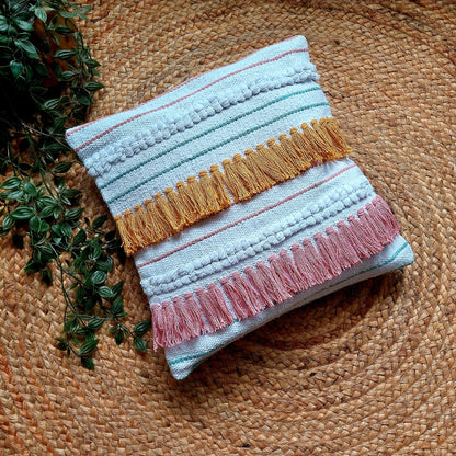 Handwoven white cushion cover with pink and mustard yellow tassels, placed on a round brown platform with a green plant in the background.