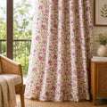 Floral curtain panel hanging in a sunlit room, with a cream background and pink, purple, and yellow flower print. The curtain is styled in a home setting next to a wooden chair and side table.