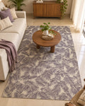 A full view of a muted purple and cream botanical leaf patterned flatweave cotton area rug styled in a modern living room with a light sofa and wooden coffee table.