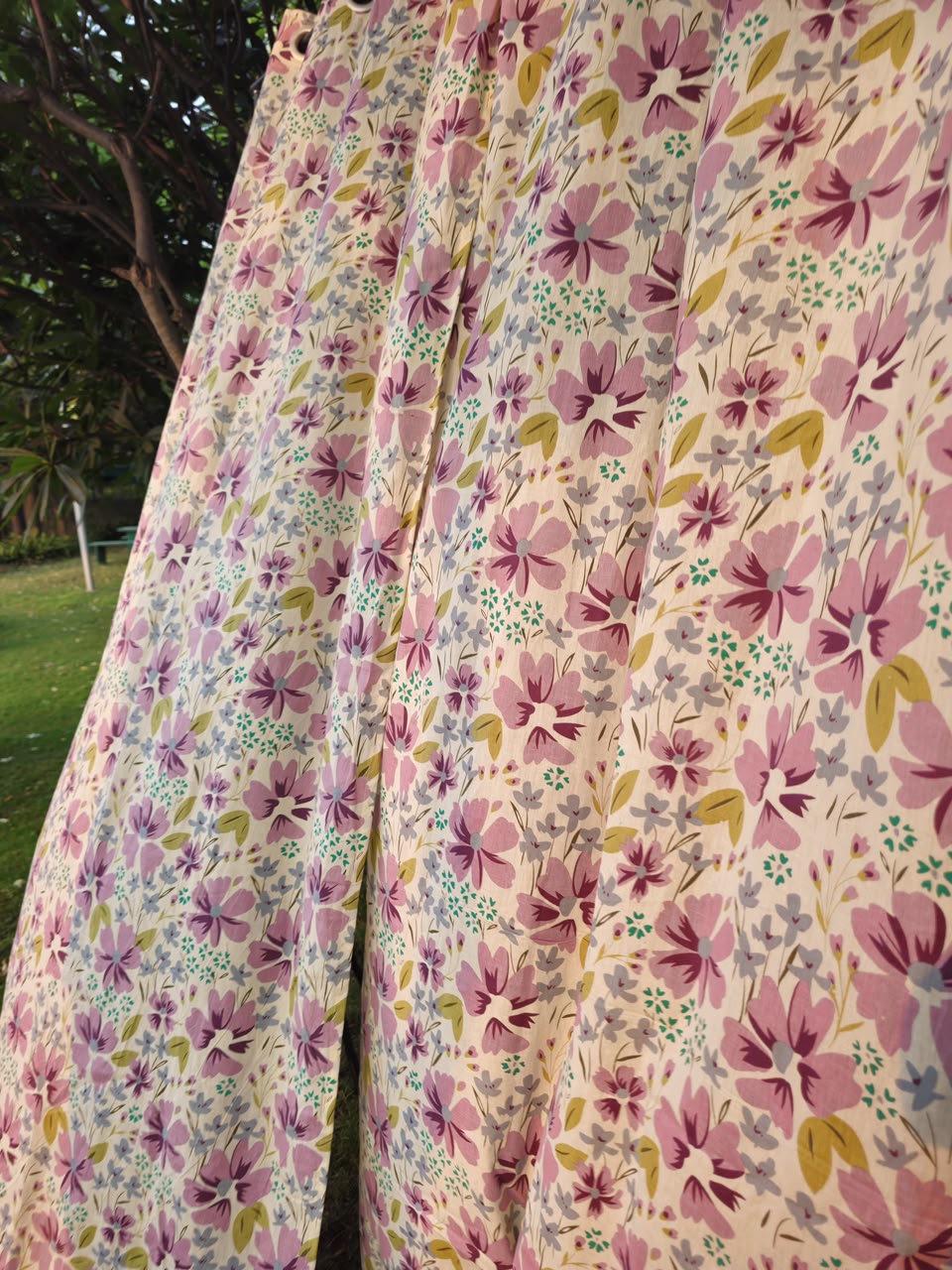 A close-up of a floral fabric, likely a curtain or drape, featuring pink and purple flowers with green and yellow accents on a light cream background. The fabric is shown hanging outdoors with a blurred green lawn and tree trunk visible in the background.