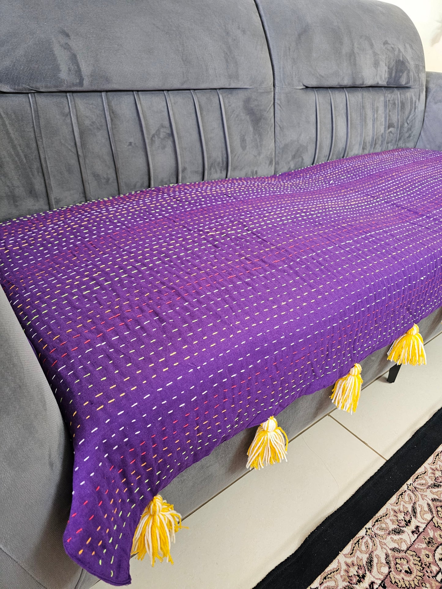 Purple textured blanket with yellow tassels draped over a gray sofa.