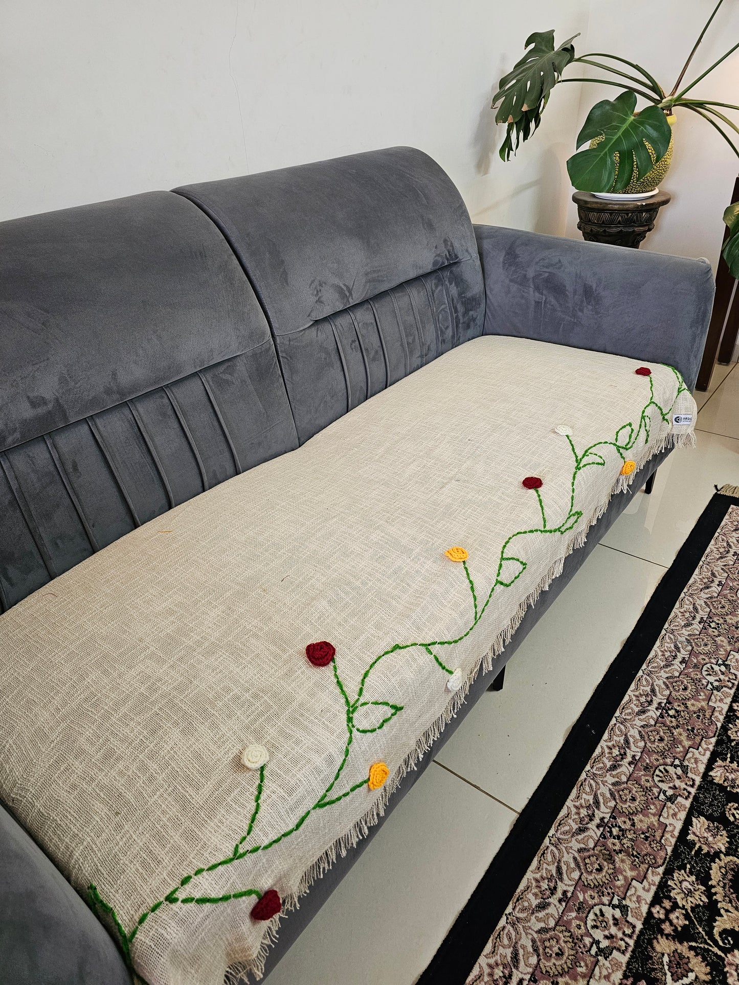 Gray sofa with a decorative white quilt featuring green vines and red flowers on a light-colored floor.