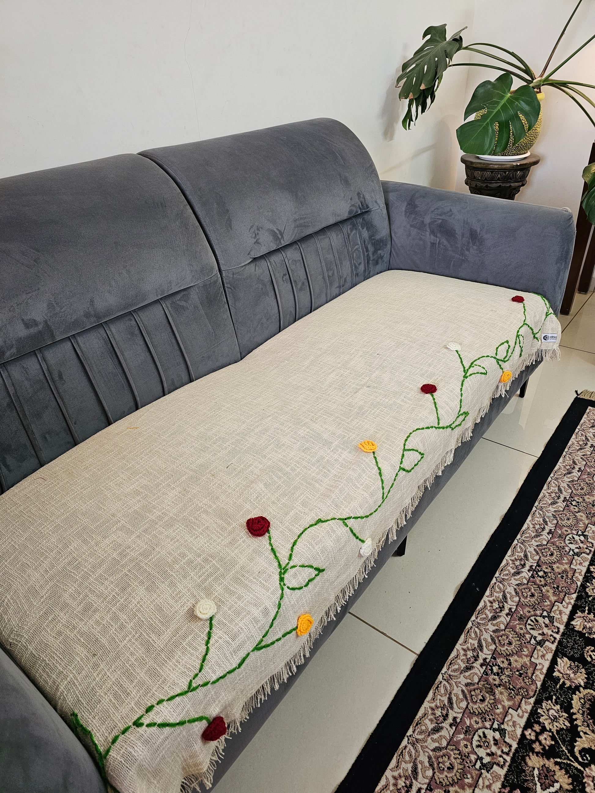 Gray sofa with a decorative white quilt featuring green vines and red flowers on a light-colored floor.