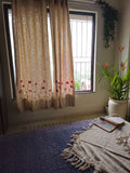 A pair of textured beige curtains with red floral embroidery near the bottom, hanging on a window in a room with a blue patterned rug and a potted plant.