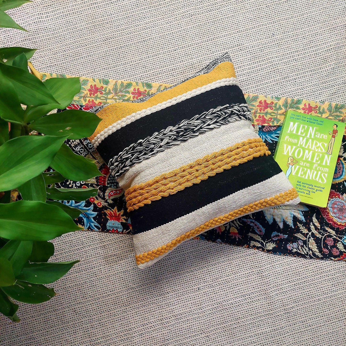 A cushion cover with a vibrant yellow base and striking black tufted patterns, placed on a patterned fabric, with a book and a potted plant in the background.