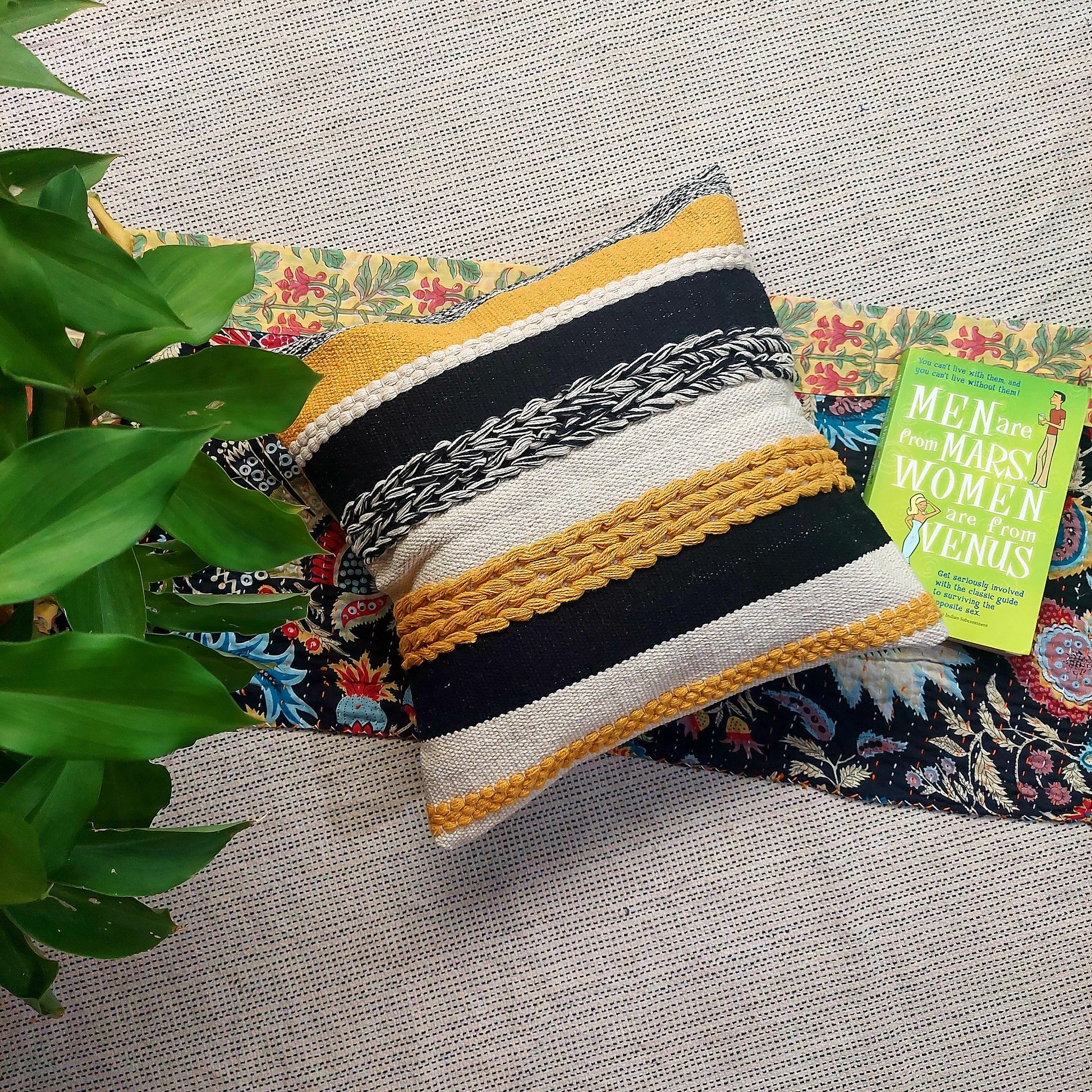 A cushion cover with a vibrant yellow base and striking black tufted patterns, placed on a patterned fabric, with a book and a potted plant in the background.