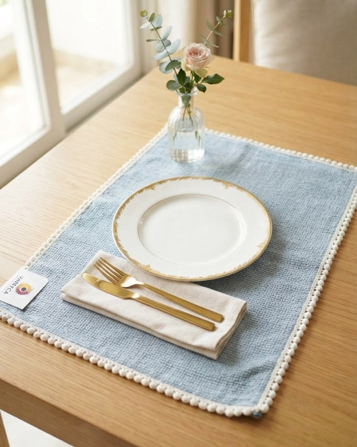 Soft blue striped cotton placemat featuring a delicate pom-pom trim border styled on a natural wooden dining table.