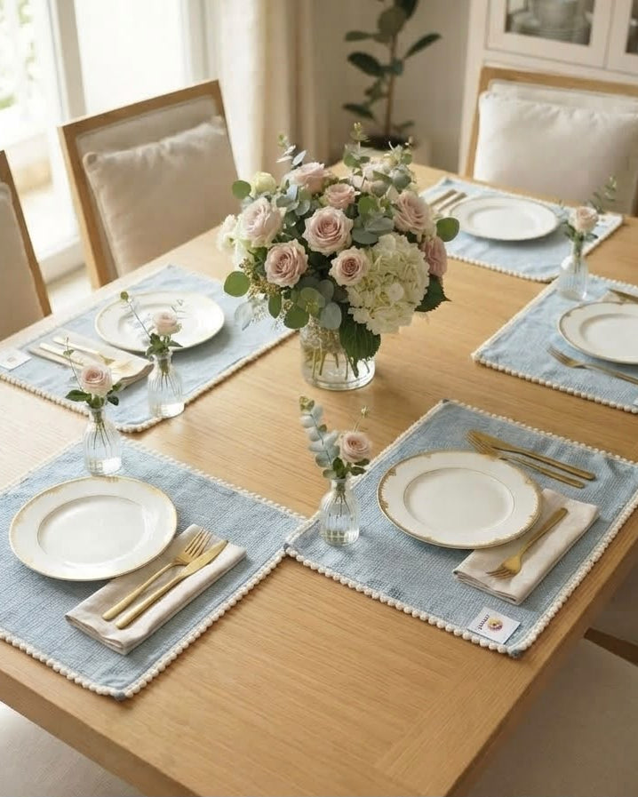 Soft blue striped cotton placemat featuring a delicate pom-pom trim border styled on a natural wooden dining table.