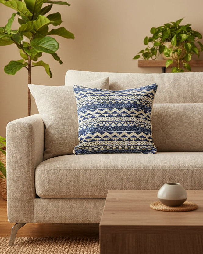 Beige sofa with a blue patterned pillow in a living room setting with plants and a coffee table.