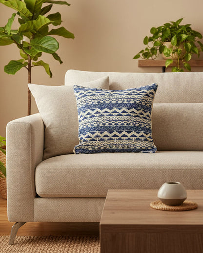 Beige sofa with a blue patterned pillow in a living room setting with plants and a coffee table.