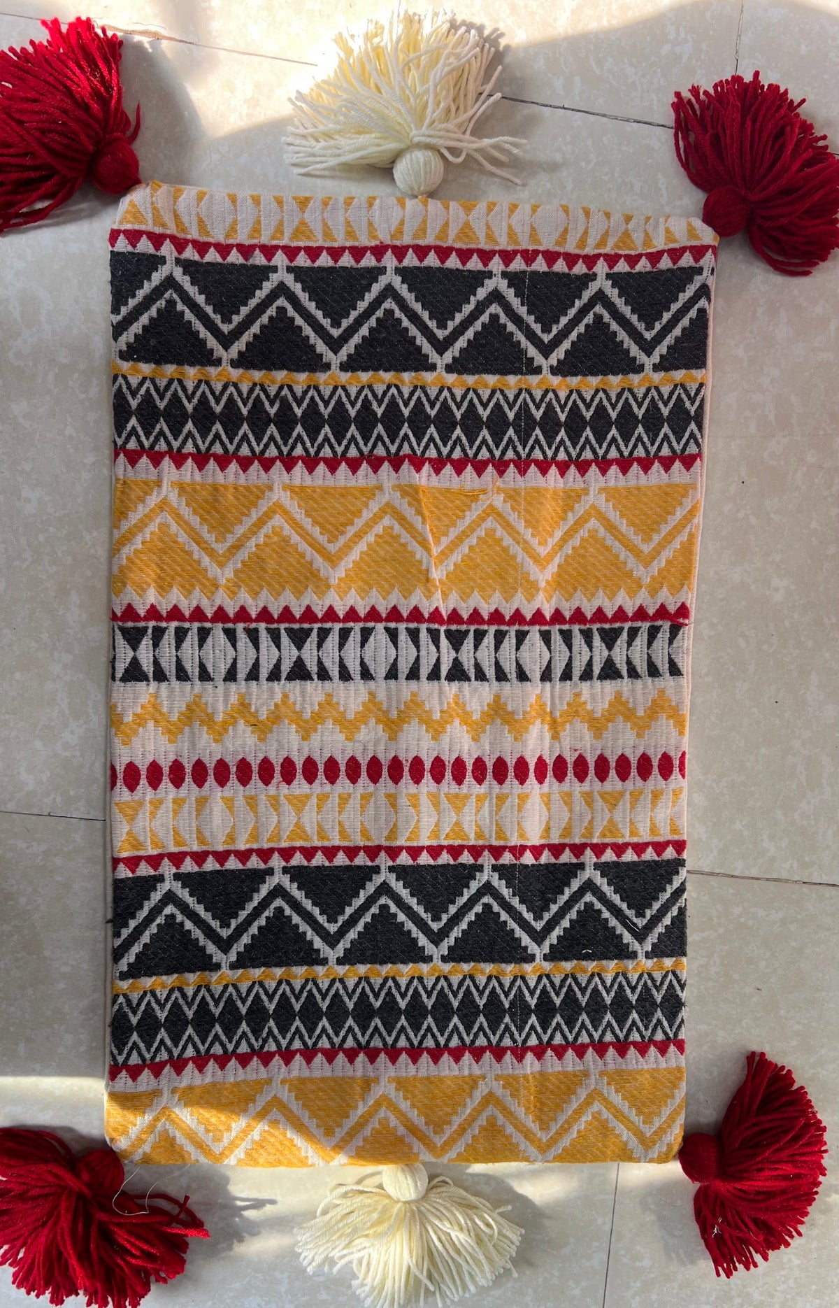 Rectangular boho-style cotton cushion cover featuring a colorful Aztec geometric pattern in yellow, black, and red, complete with red corner tassels and white center tassels, laid flat on a light tiled surface.