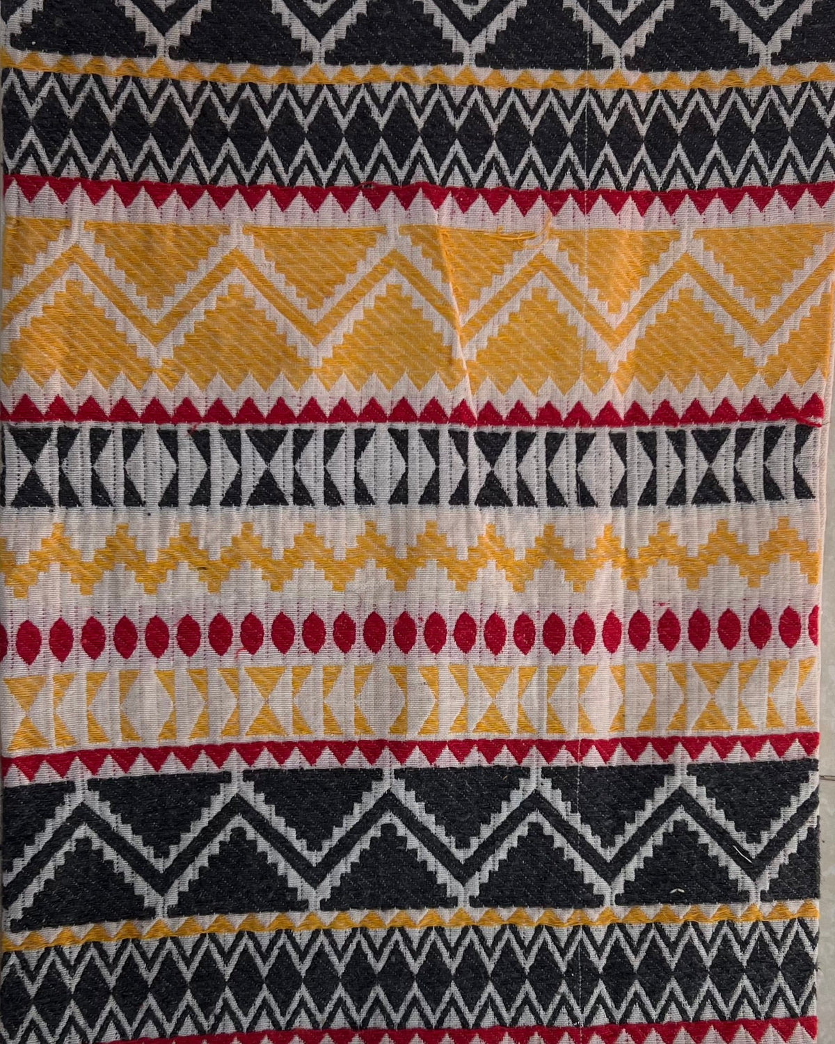 Rectangular boho-style cotton cushion cover featuring a colorful Aztec geometric pattern in yellow, black, and red, complete with red corner tassels and white center tassels, laid flat on a light tiled surface.