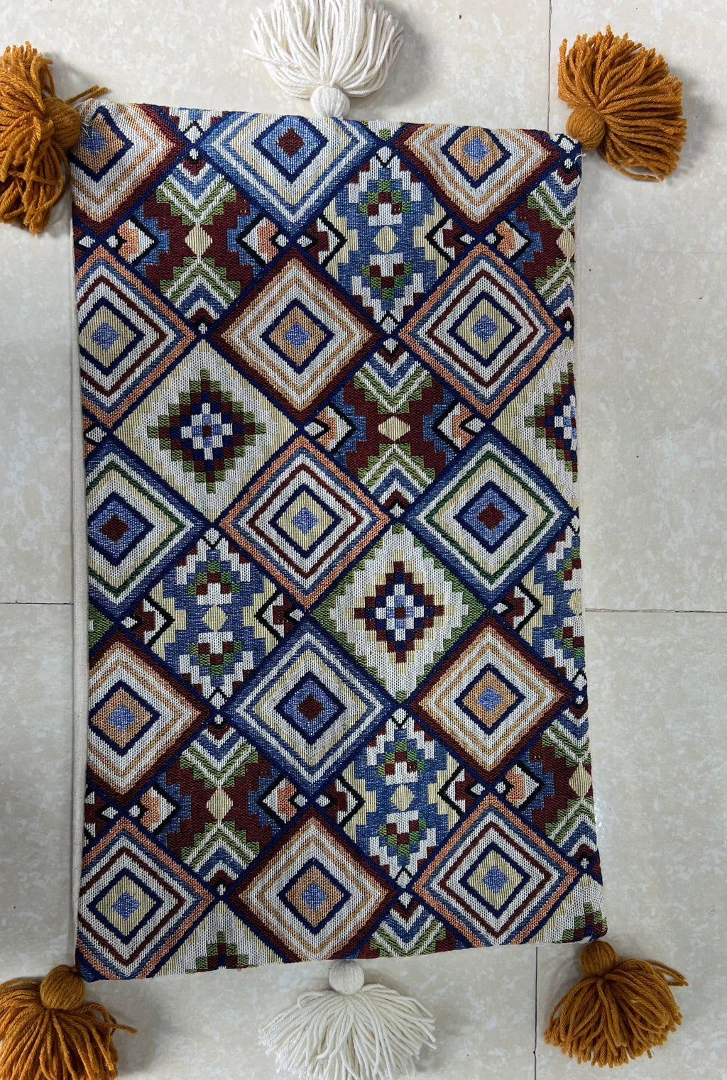 Rectangular boho-style cotton cushion cover featuring a colorful geometric diamond patchwork pattern in blue, red, and brown, complete with mustard corner tassels and white center tassels, laid flat on a light surface.