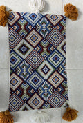 Rectangular boho-style cotton cushion cover featuring a colorful geometric diamond patchwork pattern in blue, red, and brown, complete with mustard corner tassels and white center tassels, laid flat on a light surface.