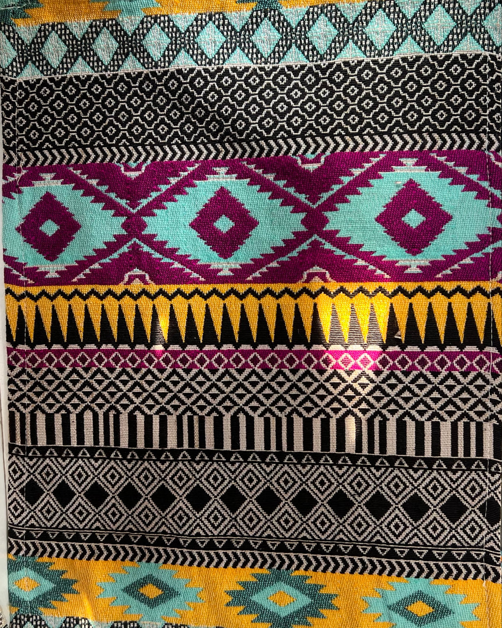 Vibrant boho-style woven table placemat featuring colorful geometric Aztec patterns in turquoise, magenta, yellow, and black, laid flat on a surface.