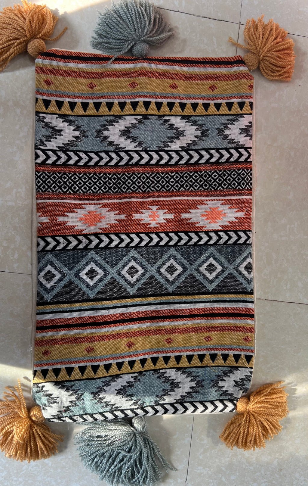 Rectangular boho-style cotton cushion cover featuring a colorful tribal geometric pattern in yellow, grey, and orange, complete with mustard corner tassels and grey center tassels, laid flat on a light surface.