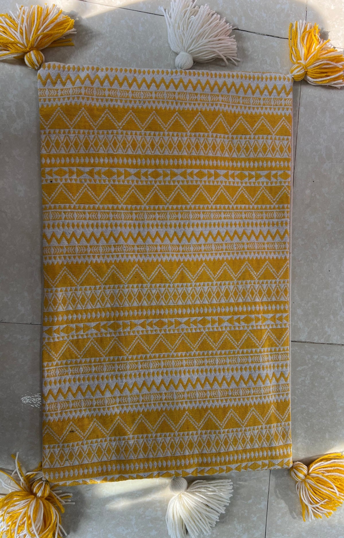 Rectangular boho-style cotton cushion cover featuring a geometric zigzag pattern in mustard yellow and white, complete with yellow-and-white corner tassels and white center tassels, laid flat on a light surface.