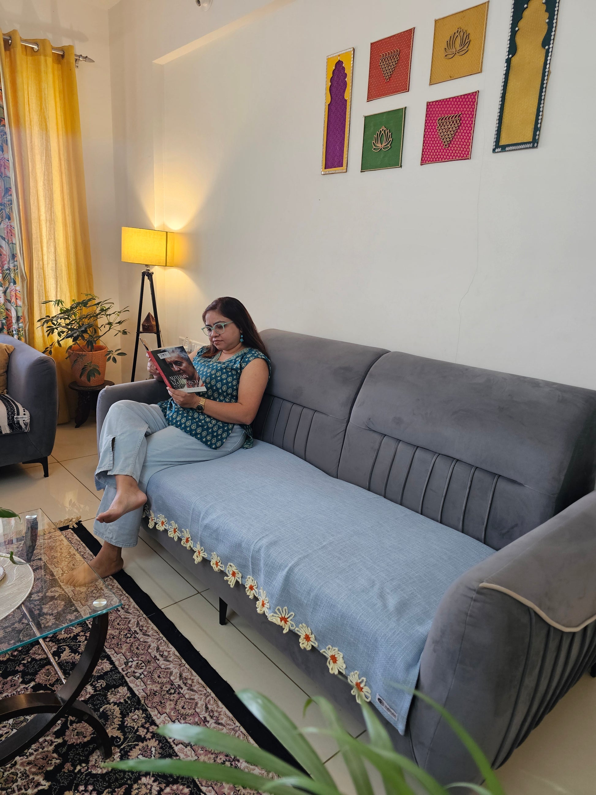 Woman sitting on a gray sofa with a blue cushion, reading a book in a room with colorful decorations on the wall.