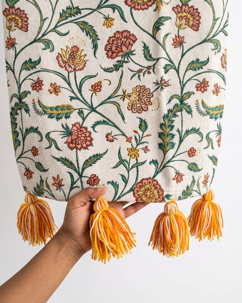 Close-up view of an off-white botanical floral printed cotton sofa cover showcasing intricate woven vine patterns and hand-tied orange and yellow yarn tassels.   