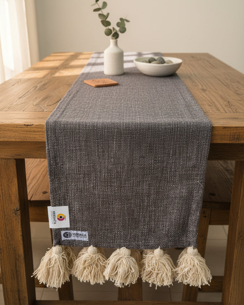 Charcoal grey jute-cotton handloom table runner featuring chunky off-white woolen hanging tassels and Vasaca brand labels.