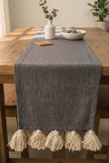 Charcoal grey jute-cotton handloom table runner featuring chunky off-white woolen hanging tassels and Vasaca brand labels.