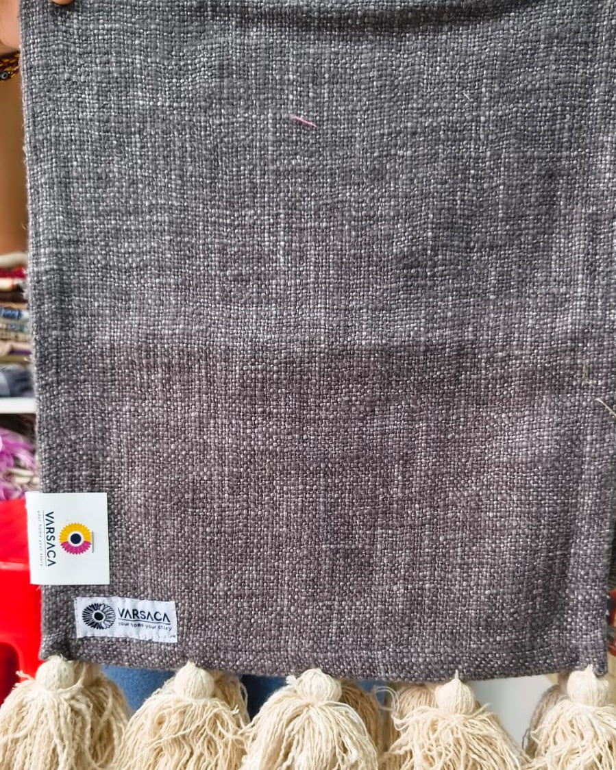 Charcoal grey jute-cotton handloom table runner featuring chunky off-white woolen hanging tassels and Vasaca brand labels.