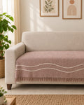 Dusty rose handwoven cotton wool sofa throw cover with off-white wavy rope lines and fringe hem draped over a cream sofa.