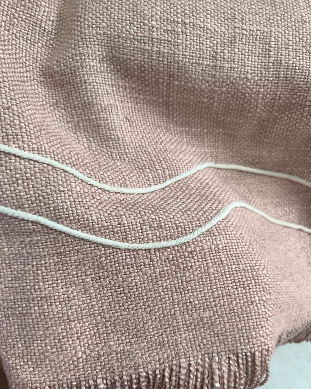 Dusty rose handwoven cotton wool sofa throw cover with off-white wavy rope lines and fringe hem draped over a cream sofa.