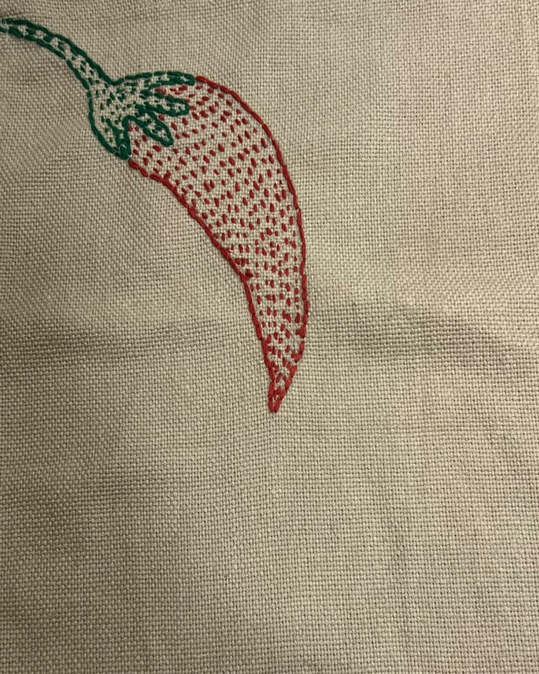 Hand-embroidered red chili pepper motif with green stem on a natural textured burlap-style cotton placemat.