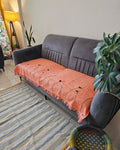 Peach coral hand-embroidered jute cotton sofa seating cover with tassels laid across a dark gray velvet sofa in a modern living room setting.