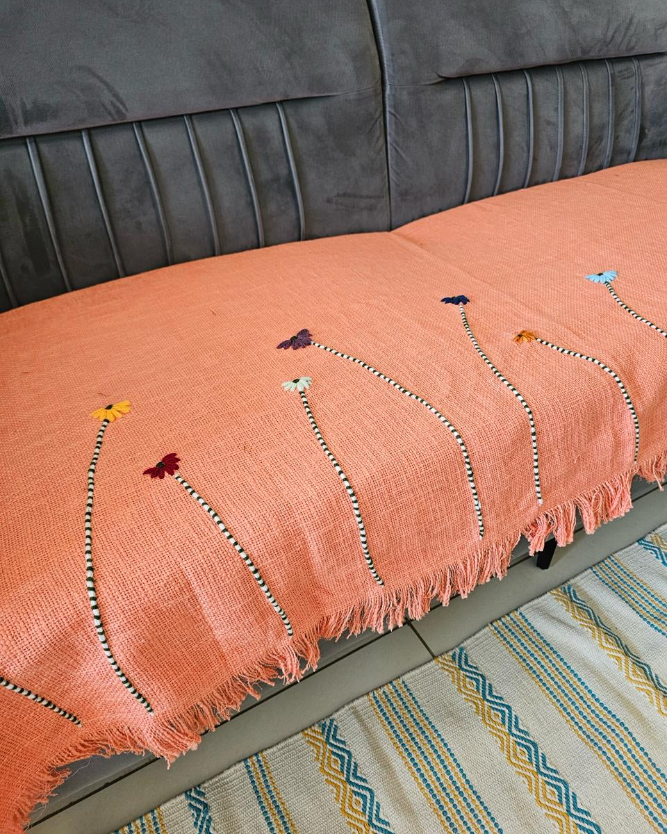 Peach coral hand-embroidered jute cotton sofa seating cover with tassels laid across a dark gray velvet sofa in a modern living room setting.